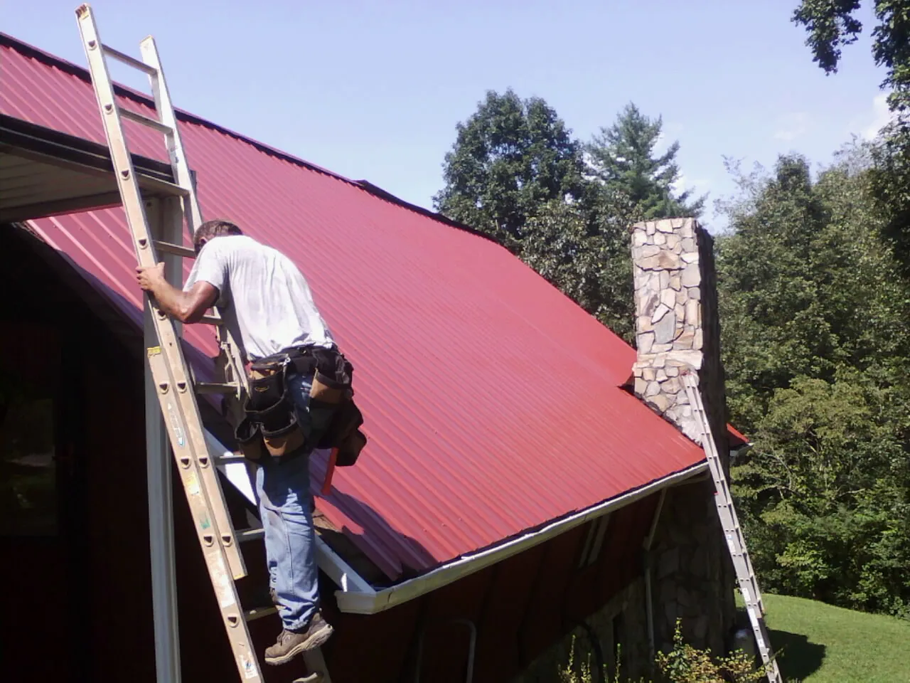 Roofer on a ladder beside a red metal roof and stone chimney — Soffit & Fascia Repair by Roofing Brothers in Long Island